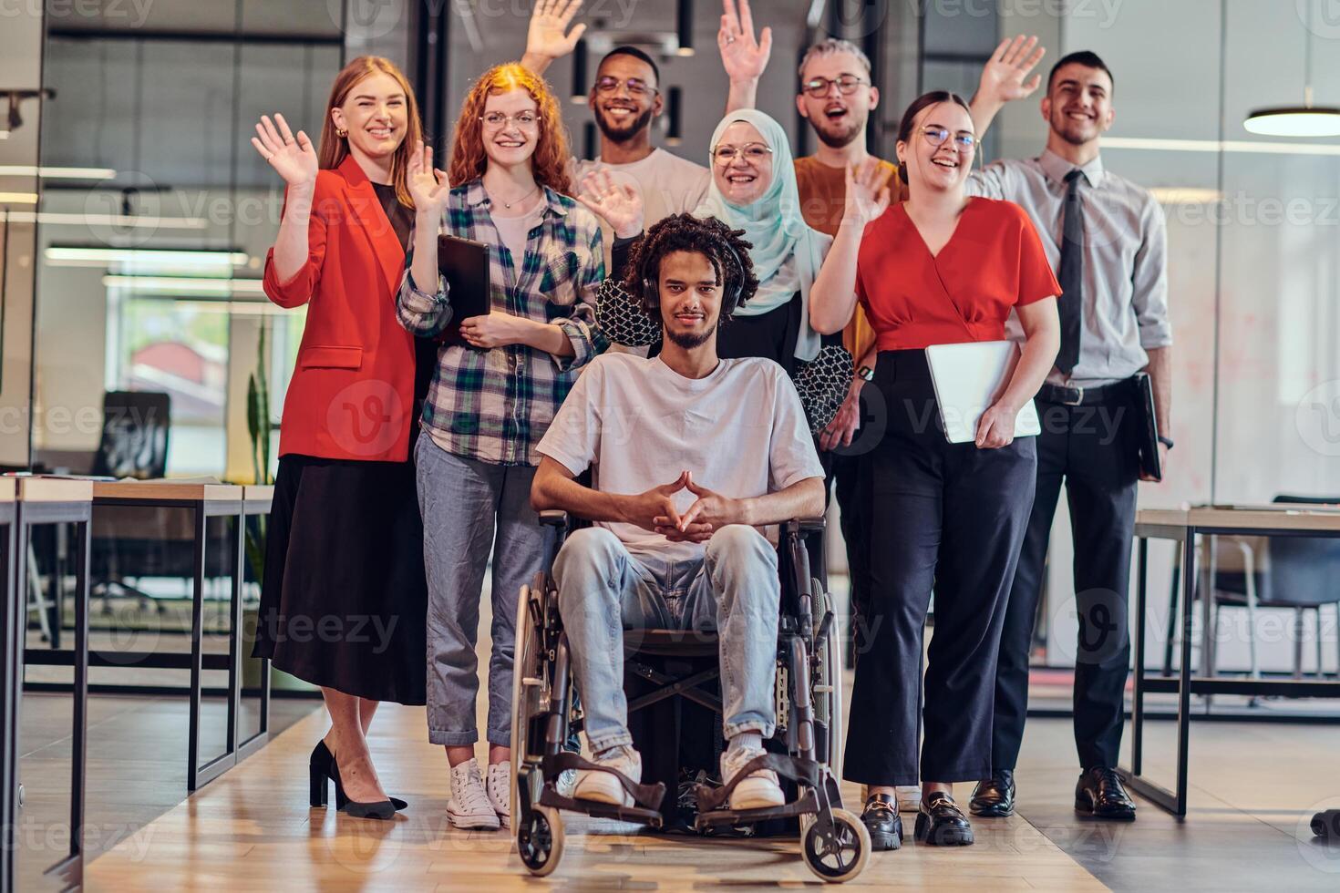 A diverse group of young business people congregates within a modern startup's glass-enclosed office, featuring inclusivity with a person in a wheelchair, an African American young man , and a hijab photo
