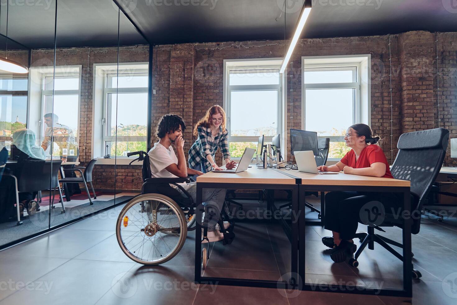 A young business group, including an African American businessman in a wheelchair, collaborates within a modern glass office, actively engaged around a computer and laptop, collectively solving photo