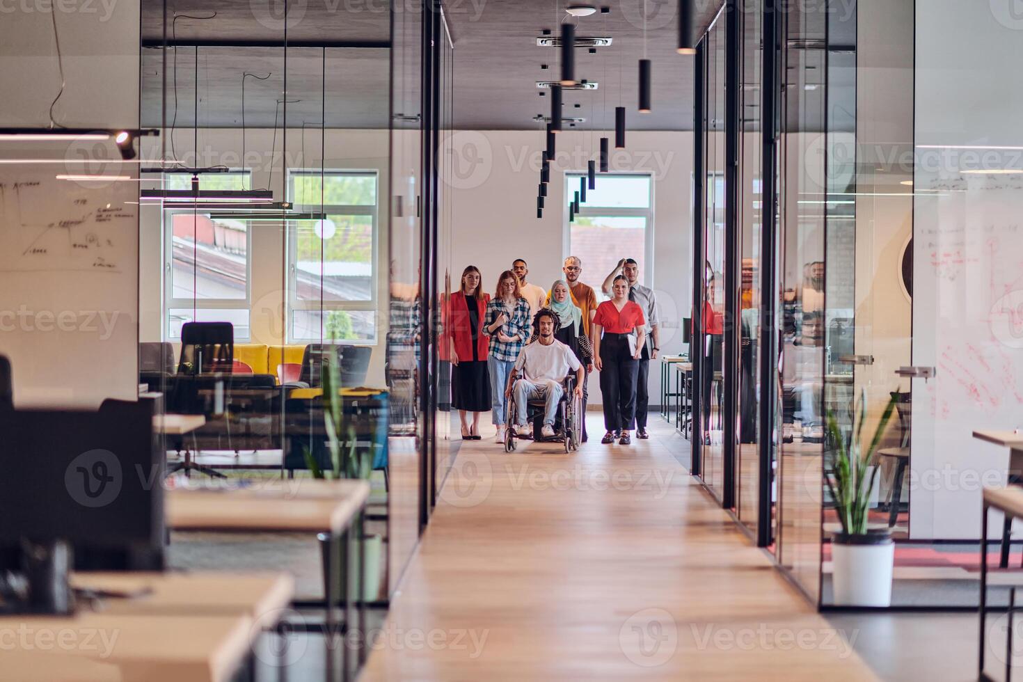 A diverse group of young business people walking a corridor in the glass-enclosed office of a modern startup, including a person in a wheelchair and a woman wearing a hijab, showing a dynamic mix of photo