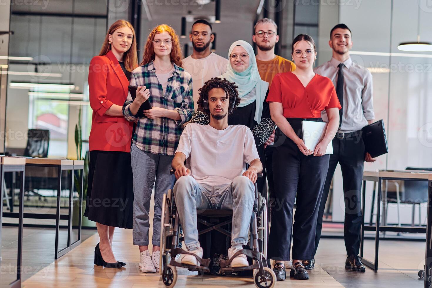 A diverse group of young business people congregates within a modern startup's glass-enclosed office, featuring inclusivity with a person in a wheelchair, an African American young man , and a hijab photo