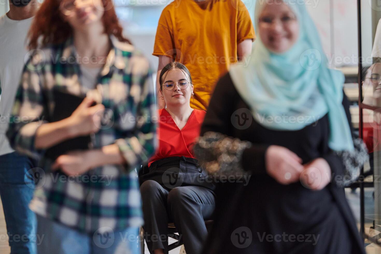 A diverse group of business people walking a corridor in the glass-enclosed office of a modern startup, including a person in a wheelchair and a woman wearing a hijab, showing a dynamic mix of photo
