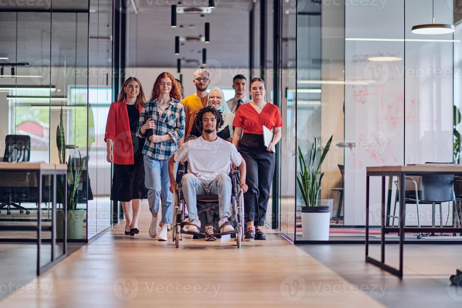 A diverse group of young business people congregates within a modern startup's glass-enclosed office, featuring inclusivity with a person in a wheelchair, an African American young man , and a hijab photo