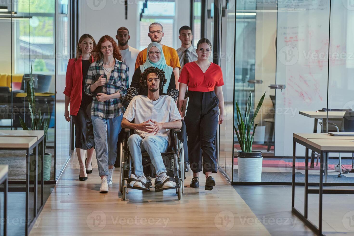 A diverse group of young business people walking a corridor in the glass-enclosed office of a modern startup, including a person in a wheelchair and a woman wearing a hijab, showing a dynamic mix of photo