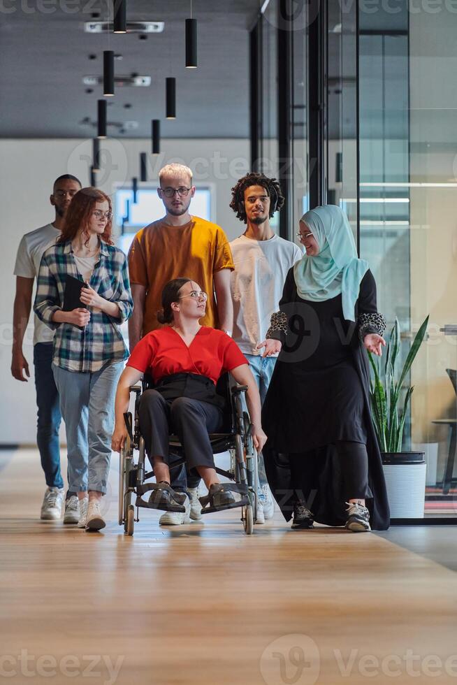 A diverse group of young business people walking a corridor in the glass-enclosed office of a modern startup, including a person in a wheelchair and a woman wearing a hijab, showing a dynamic mix of photo