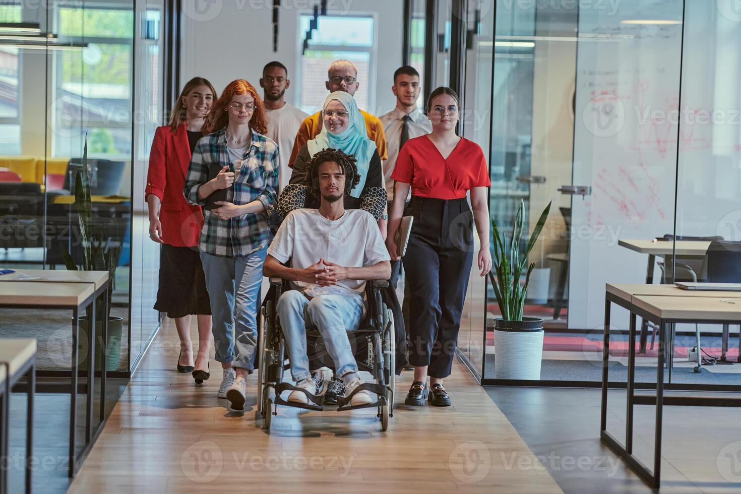 A diverse group of young business people walking a corridor in the glass-enclosed office of a modern startup, including a person in a wheelchair and a woman wearing a hijab, showing a dynamic mix of photo