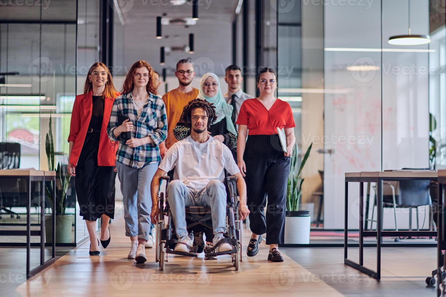 A diverse group of young business people congregates within a modern startup's glass-enclosed office, featuring inclusivity with a person in a wheelchair, an African American young man , and a hijab photo