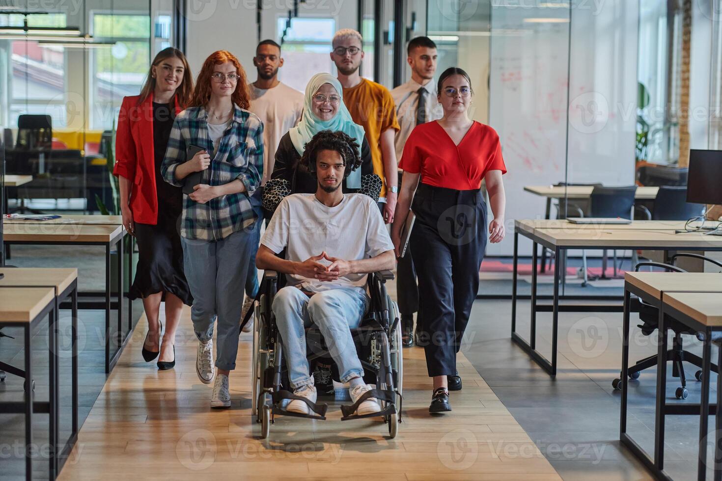 A diverse group of young business people walking a corridor in the glass-enclosed office of a modern startup, including a person in a wheelchair and a woman wearing a hijab, showing a dynamic mix of photo