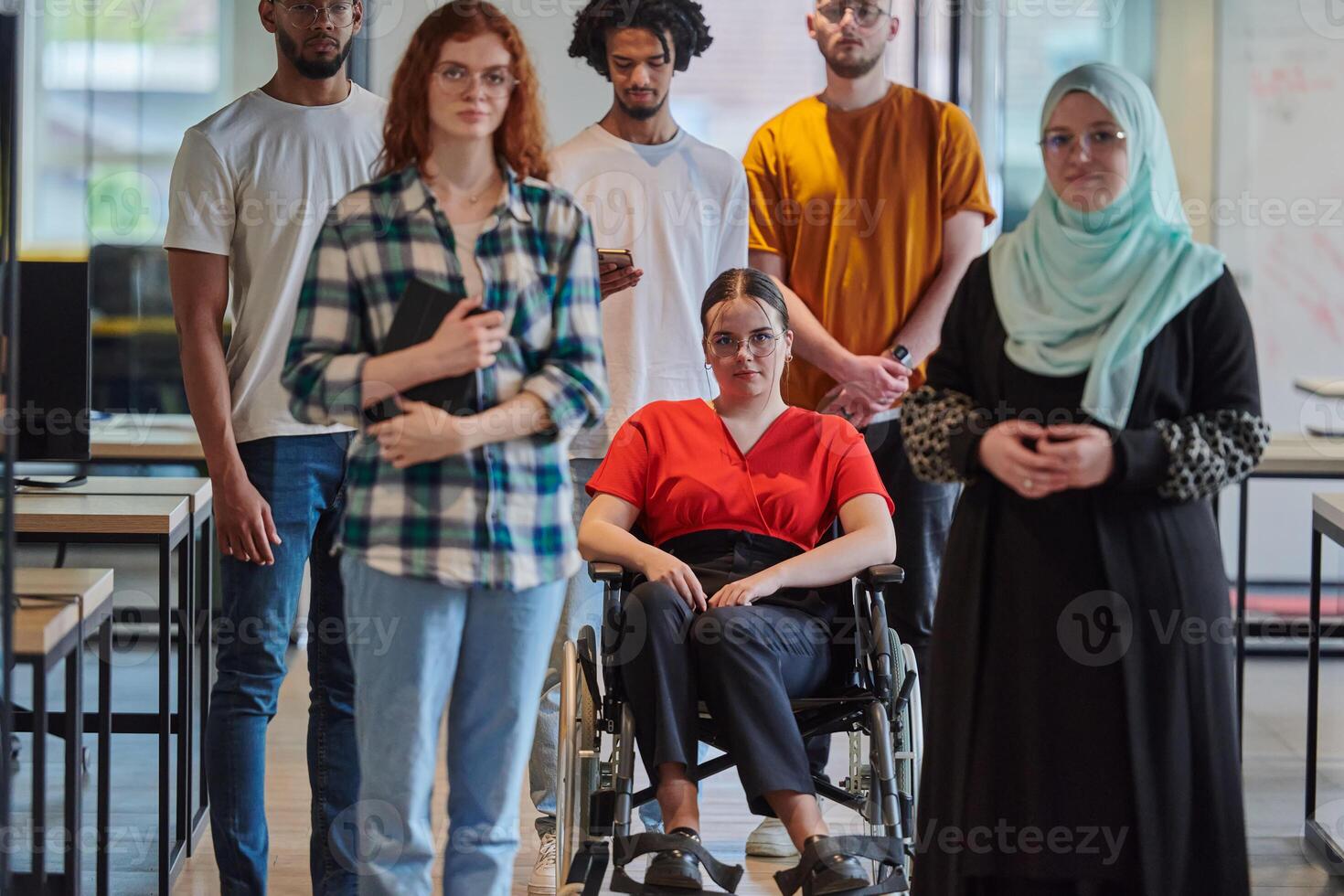 A diverse group of young business people walking a corridor in the glass-enclosed office of a modern startup, including a person in a wheelchair and a woman wearing a hijab, showing a dynamic mix of photo