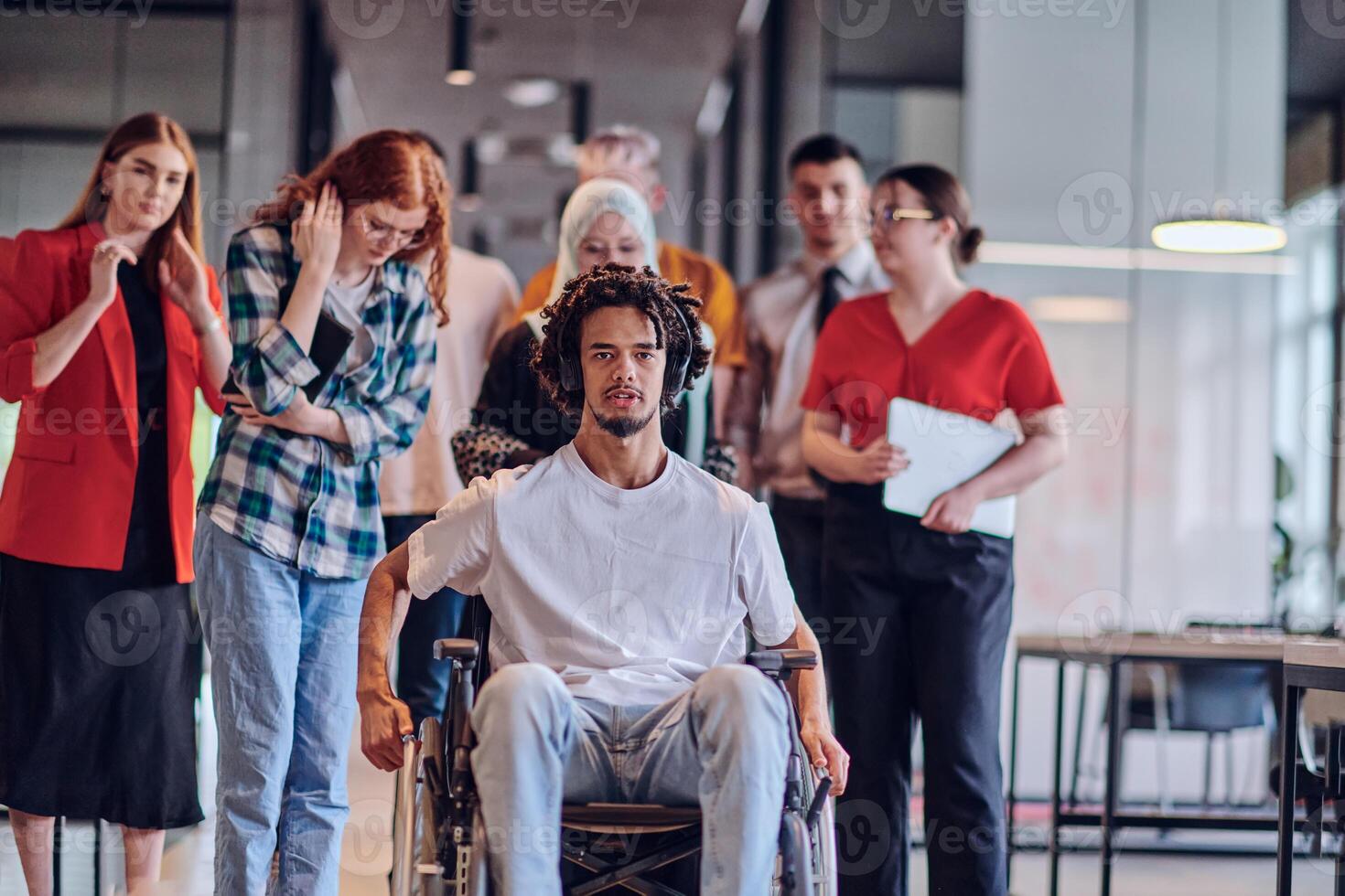 A diverse group of young business people congregates within a modern startup's glass-enclosed office, featuring inclusivity with a person in a wheelchair, an African American young man , and a hijab photo