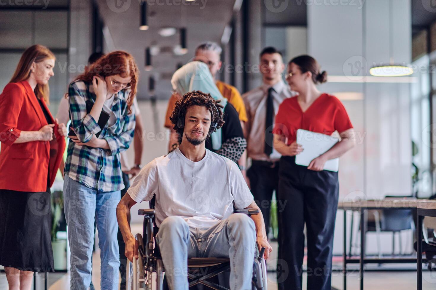 A diverse group of young business people congregates within a modern startup's glass-enclosed office, featuring inclusivity with a person in a wheelchair, an African American young man , and a hijab photo