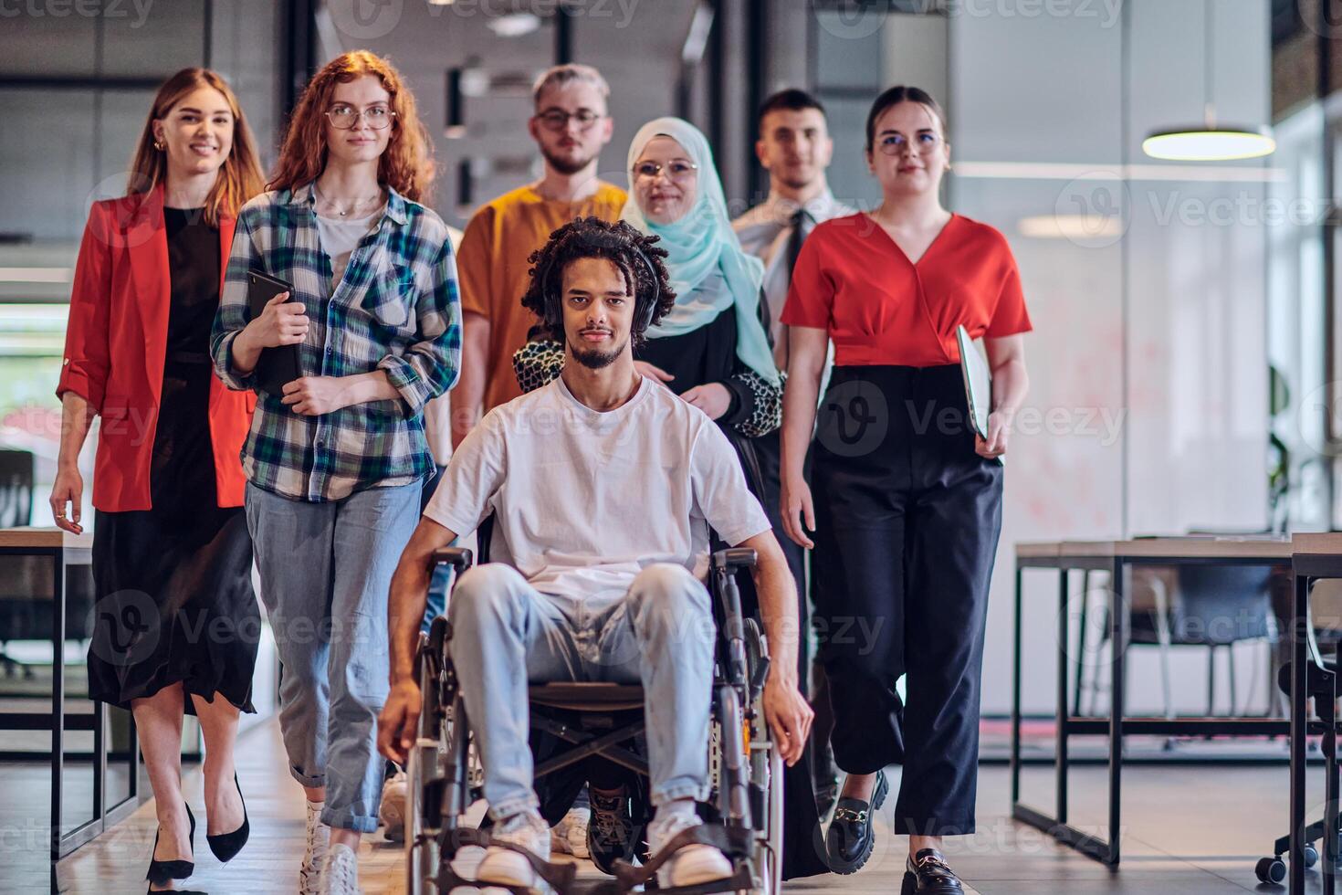A diverse group of young business people congregates within a modern startup's glass-enclosed office, featuring inclusivity with a person in a wheelchair, an African American young man , and a hijab photo