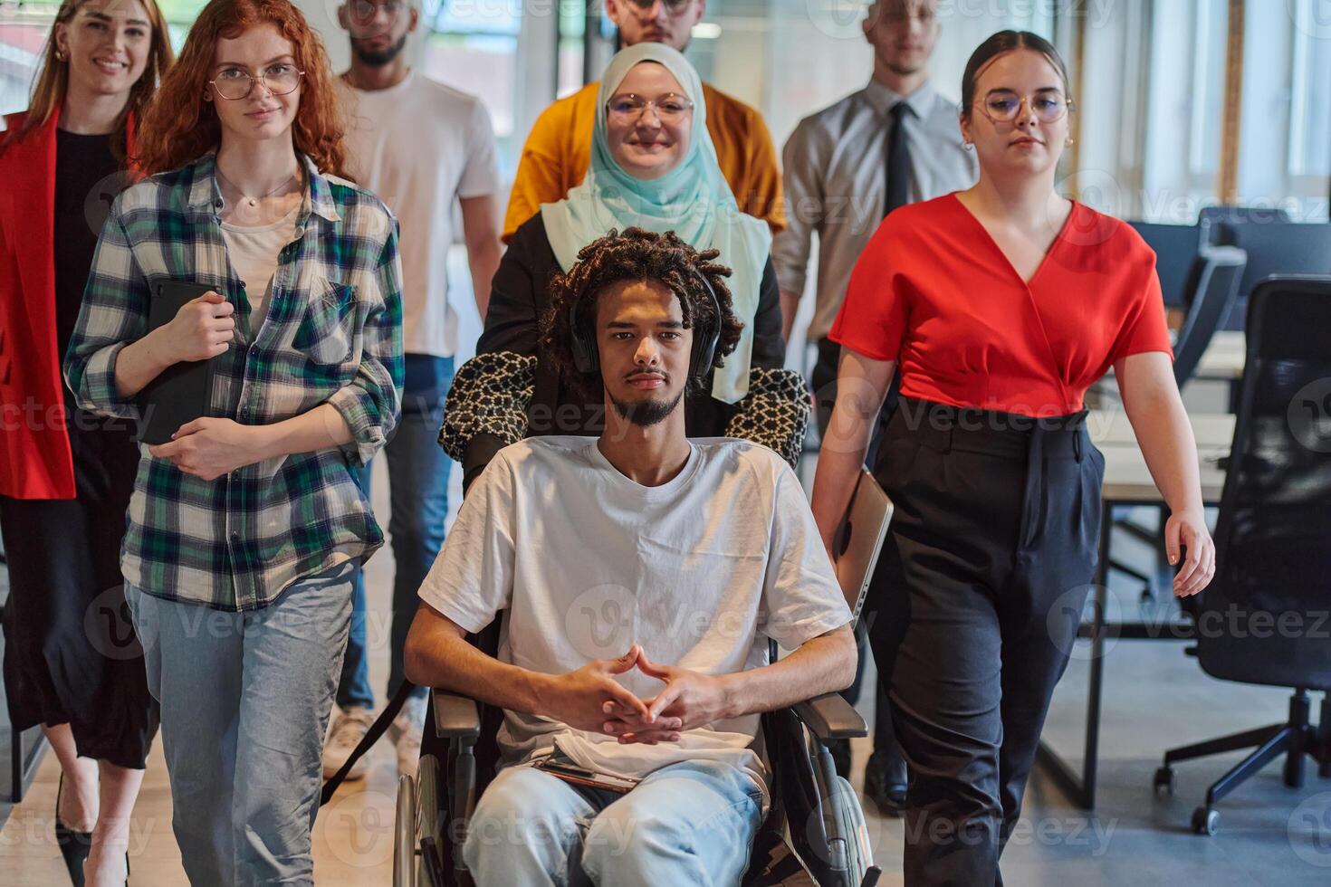 A diverse group of young business people walking a corridor in the glass-enclosed office of a modern startup, including a person in a wheelchair and a woman wearing a hijab, showing a dynamic mix of photo