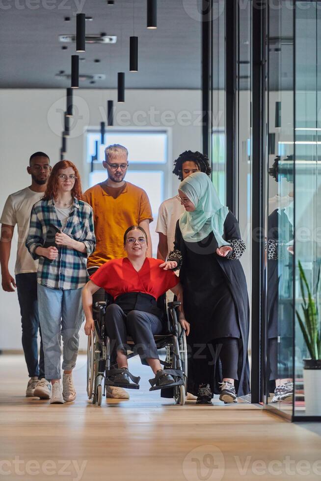 A diverse group of young business people walking a corridor in the glass-enclosed office of a modern startup, including a person in a wheelchair and a woman wearing a hijab, showing a dynamic mix of photo