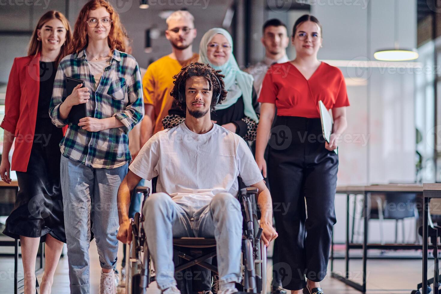 A diverse group of young business people congregates within a modern startup's glass-enclosed office, featuring inclusivity with a person in a wheelchair, an African American young man , and a hijab photo