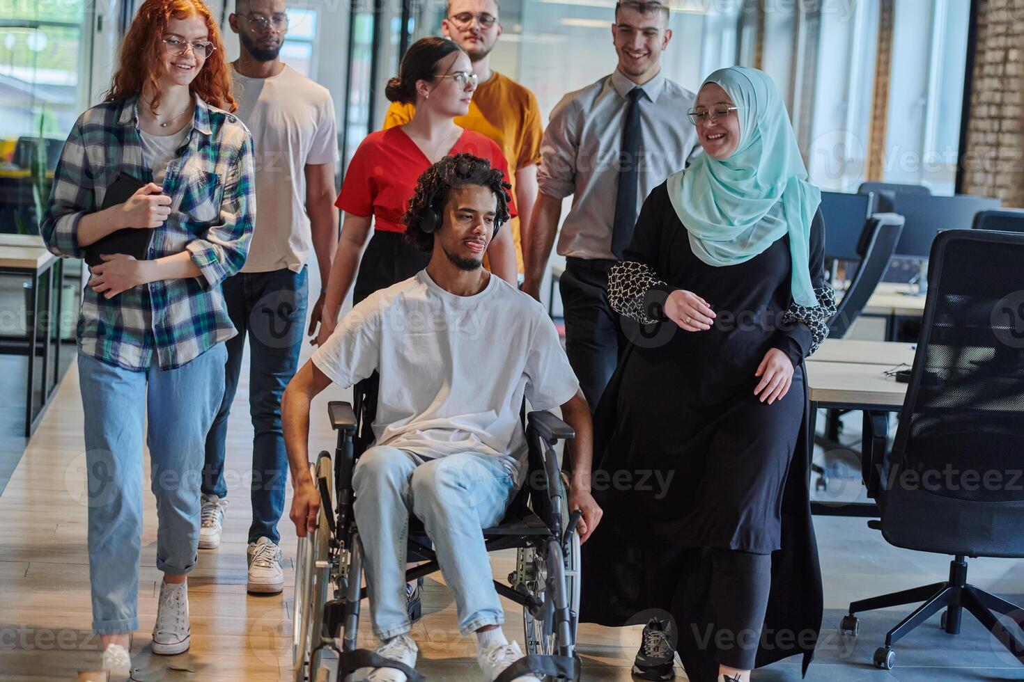 A diverse group of young business people walking a corridor in the glass-enclosed office of a modern startup, including a person in a wheelchair and a woman wearing a hijab, showing a dynamic mix of photo
