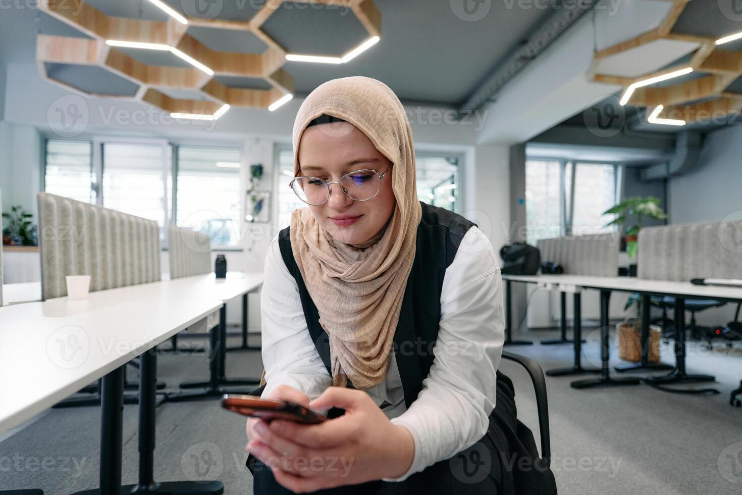 A businesswoman in a hijab using a smartphone in a modern office, epitomizing a successful and empowered professional in today's tech-savvy world photo