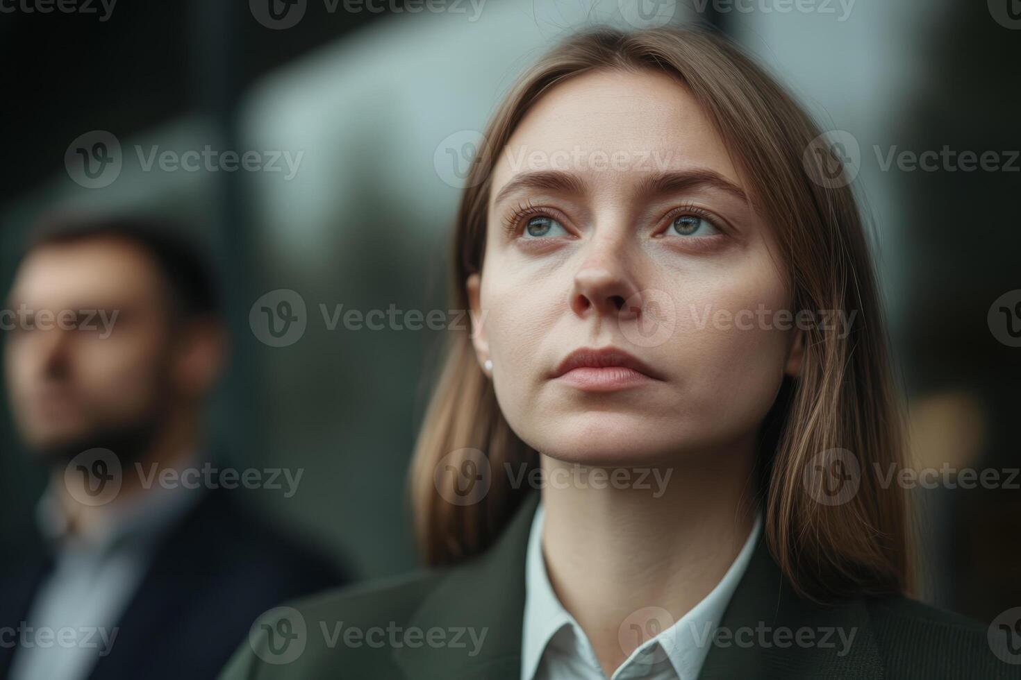 Woman with long hair and focused expression stands outdoors with a man in the background during a cloudy day photo