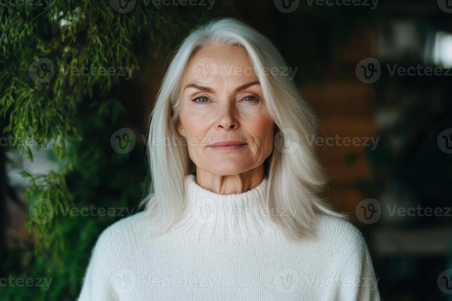 Mature woman with long gray hair in a white sweater poses in a green, indoor setting with natural light photo