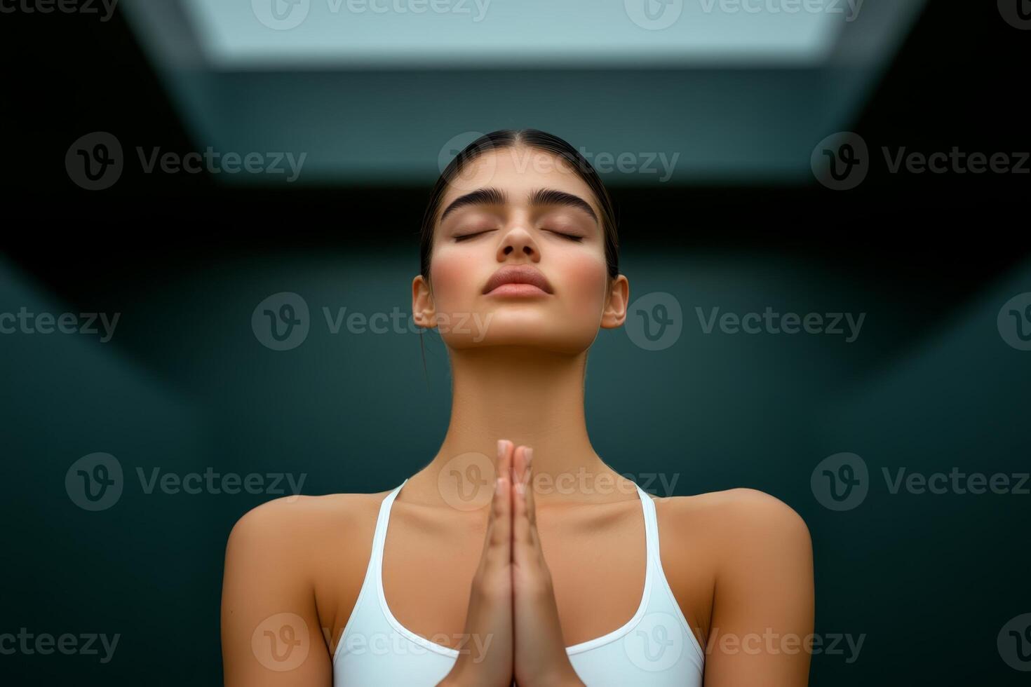 A woman is meditating with closed eyes and hands pressed together in prayer position. Soft light filters through the ceiling, creating a calm atmosphere photo