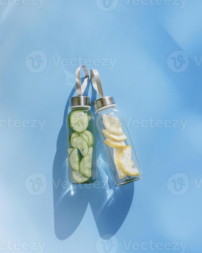 Top view two glass bottles, water drink detox with lemon and cucumber at sunlight on blue background, copy space. Infused water with slices fruit or vegetable. Aesthetic photo fresh drink