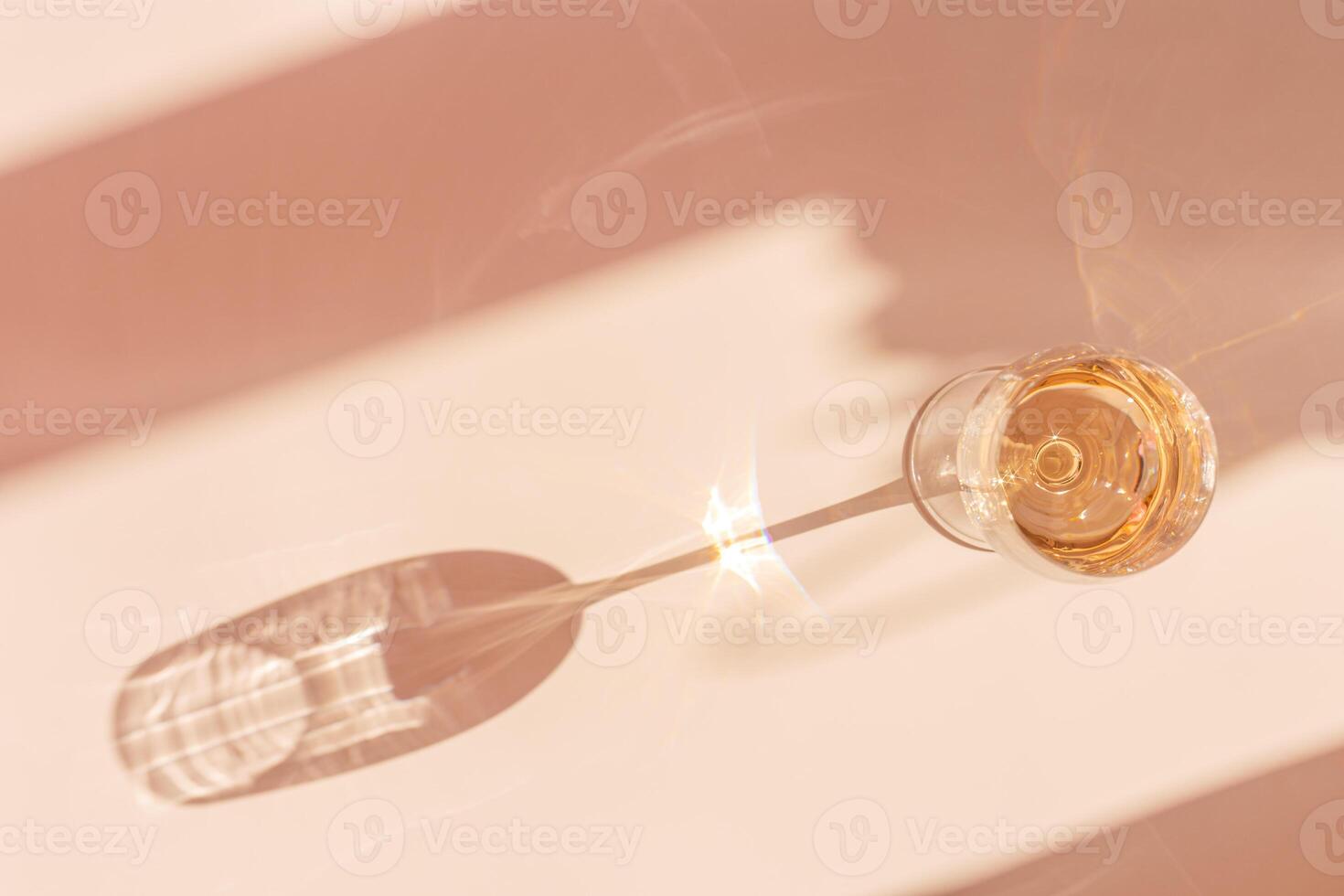 One wineglass with white wine sparkling at sunlight, beige background with dark shadow and reflection, sun flare light on table. Alcohol drink with star filter, top view, neutral pastel color photo