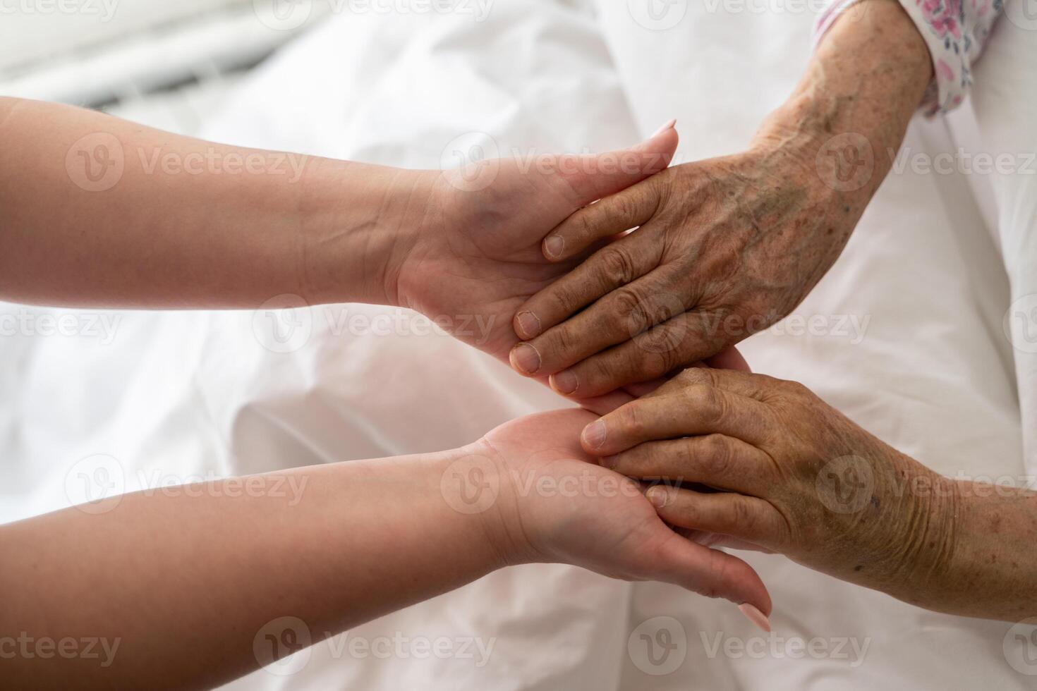 A young womans hands gently hold the hands of an elderly female patient in a hospital bed, symbolizing care, support, and compassion. photo