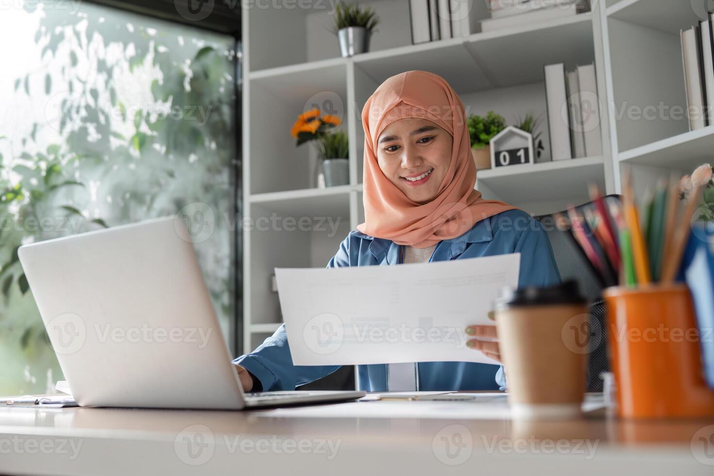 Smiling Muslim woman reviewing documents while working on a laptop in an office. photo