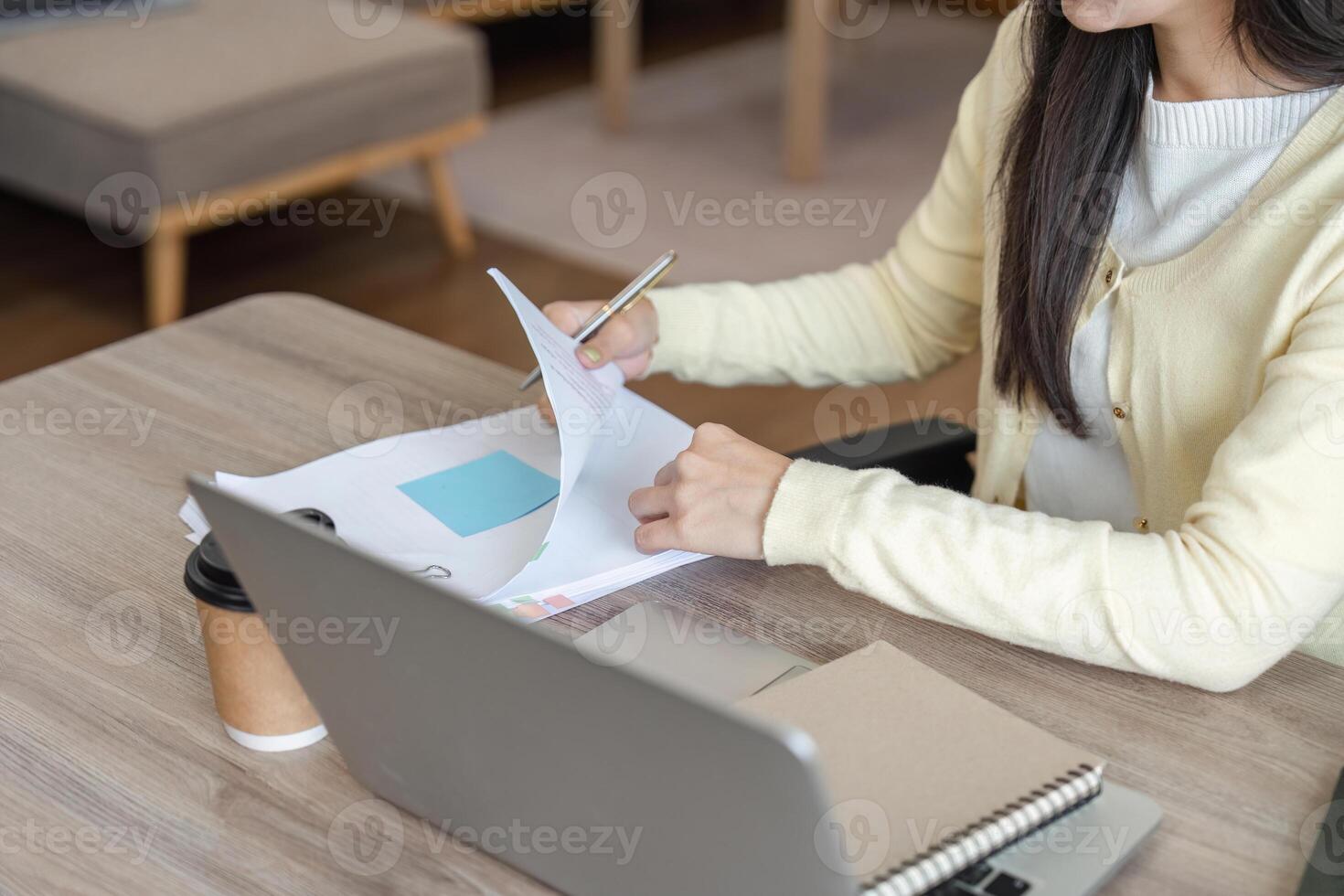 Focused Asian woman reviewing documents and taking notes while working on a laptop in a cozy home office. photo