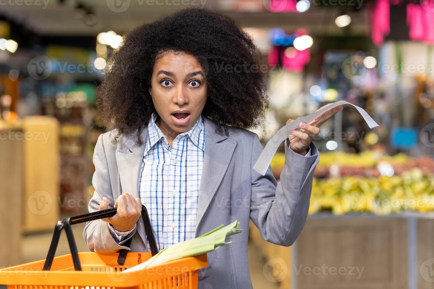 Surprised woman in business attire holding long receipt and orange shopping basket in grocery store. Expression of shock and disbelief while shopping. Concept of high prices, budgeting shopping. photo