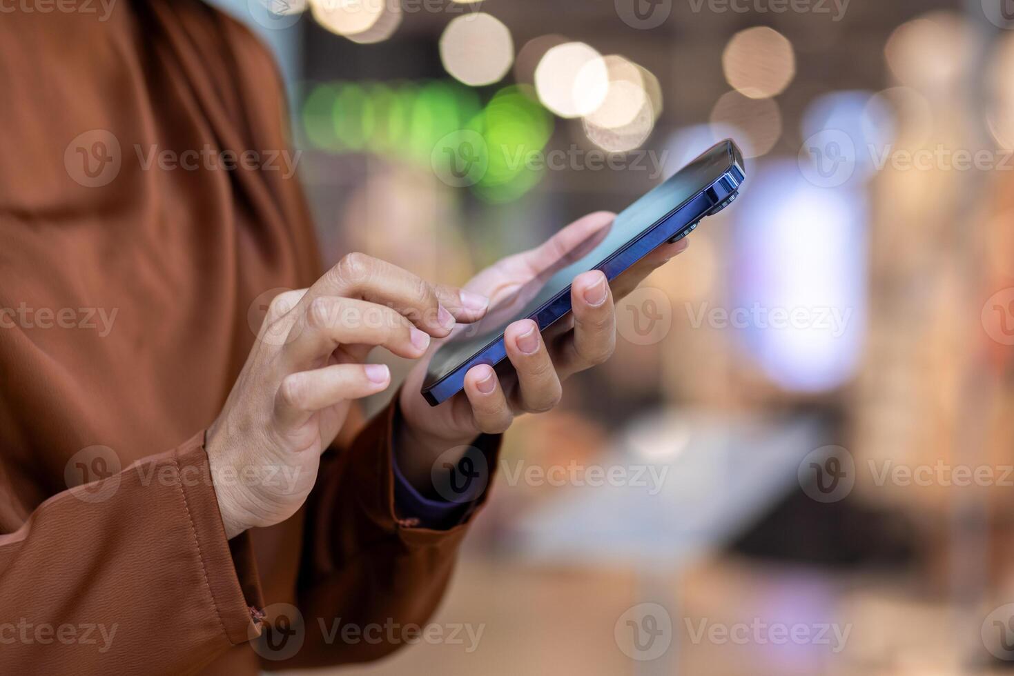 Close-up of hands interacting with a smartphone in a busy shopping mall. Background features colorful bokeh lights creating a lively, modern atmosphere, emphasizing connectivity, technology photo