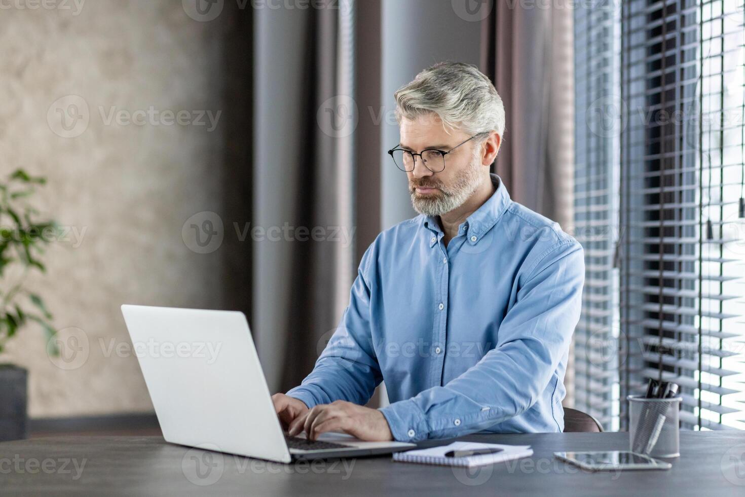 Mature professional man in blue shirt working on a laptop in a modern office setting with natural light and focus on his work. photo