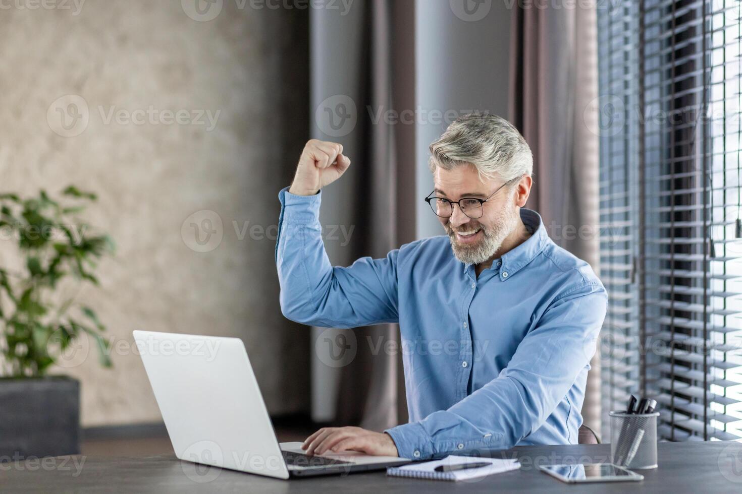 Happy businessman celebrates success with raised fist while working on laptop in a modern office. Achievement and joy in professional environment. photo