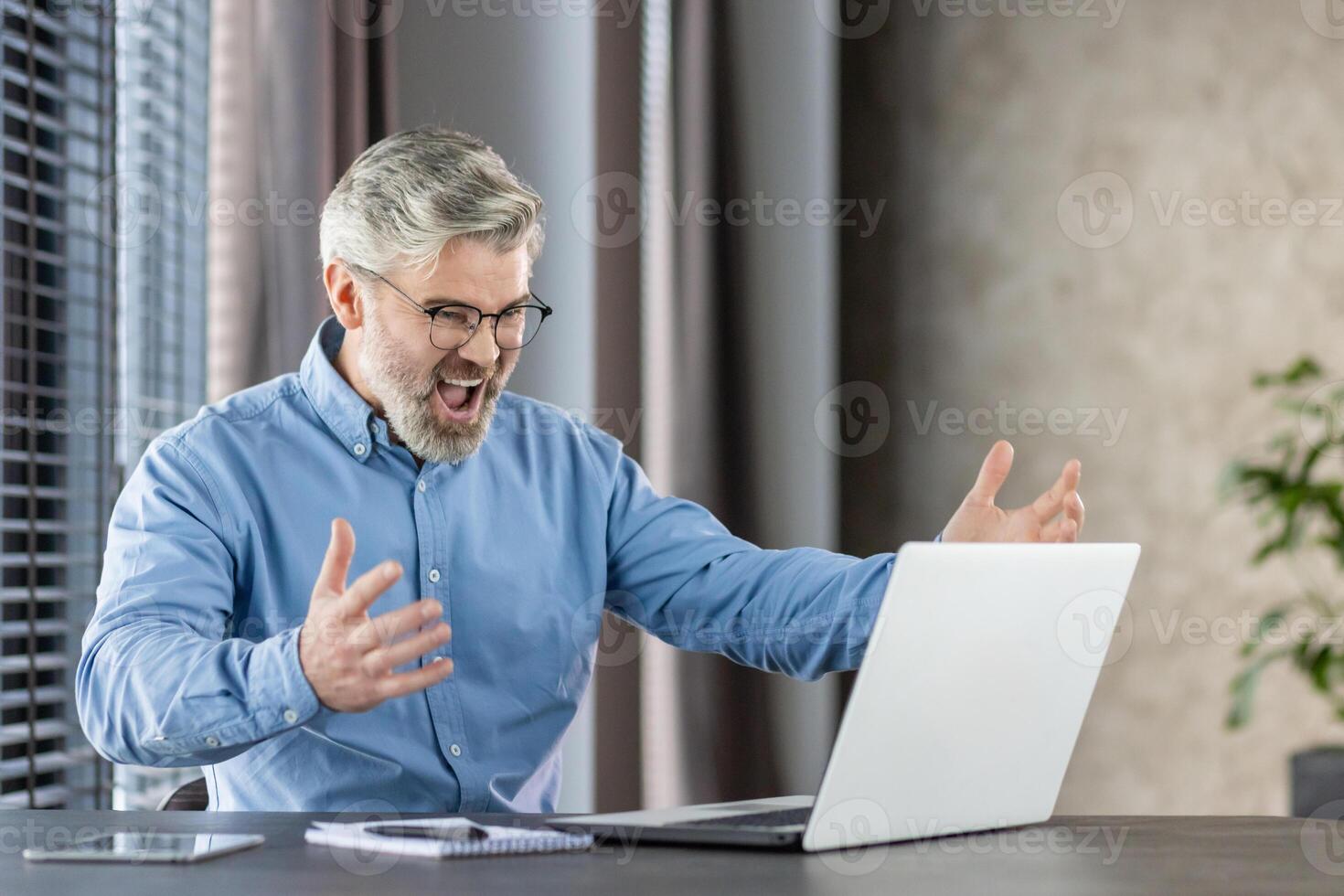 Senior businessman in a blue shirt celebrates success while working on a laptop in a modern office setting. photo