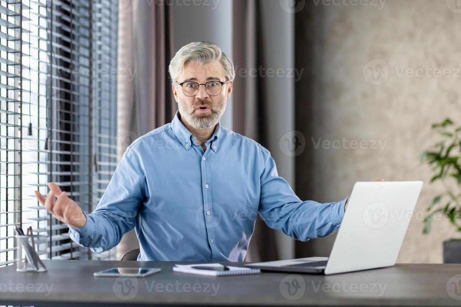 Middle-aged businessman sitting at office desk with laptop, expressing frustration and confusion. The man appears bewildered, gesturing with his hands, photo