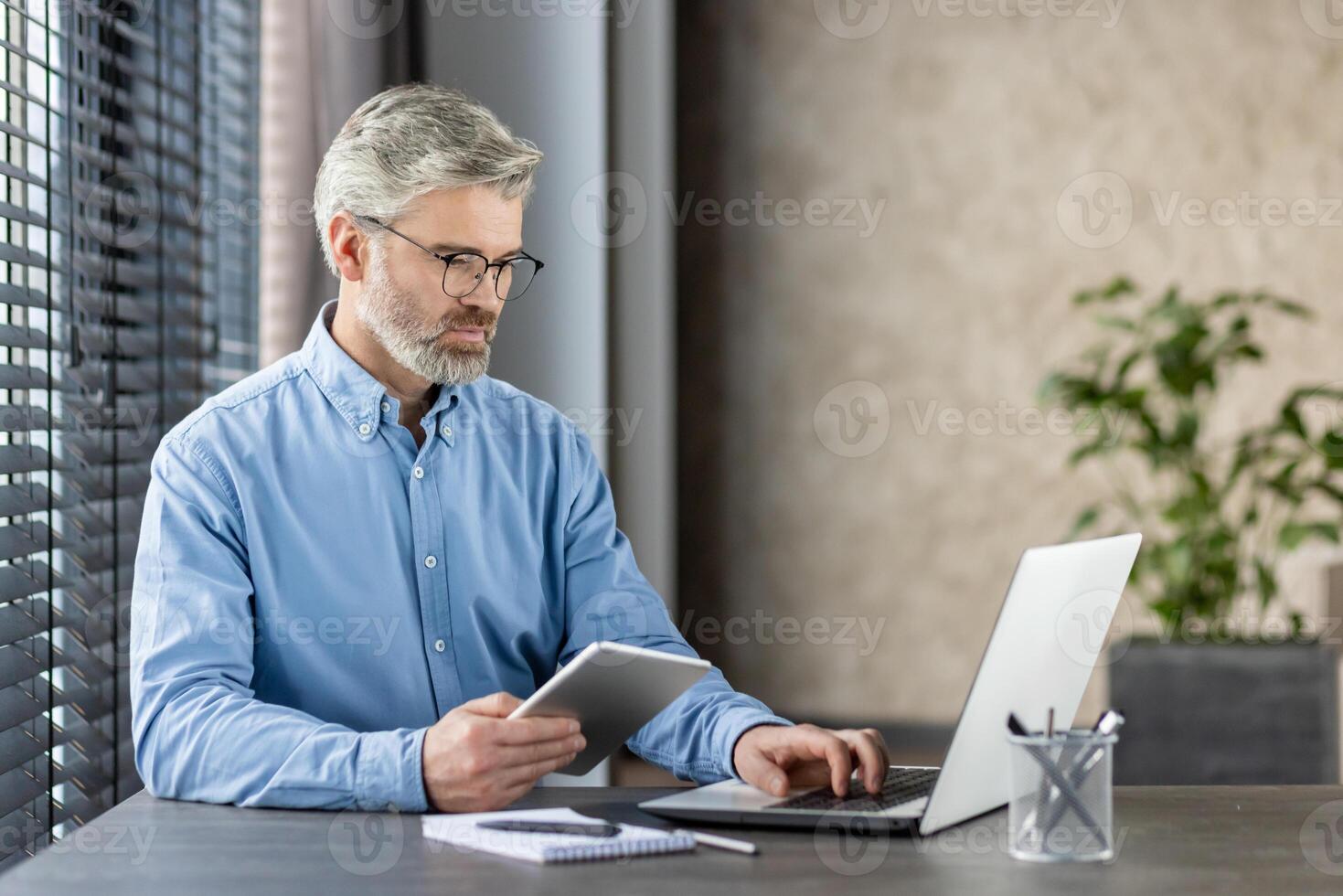 Mature office worker in a blue shirt working diligently on a laptop and tablet in a modern work environment. The scene portrays focus, concentration, and professionalism. photo