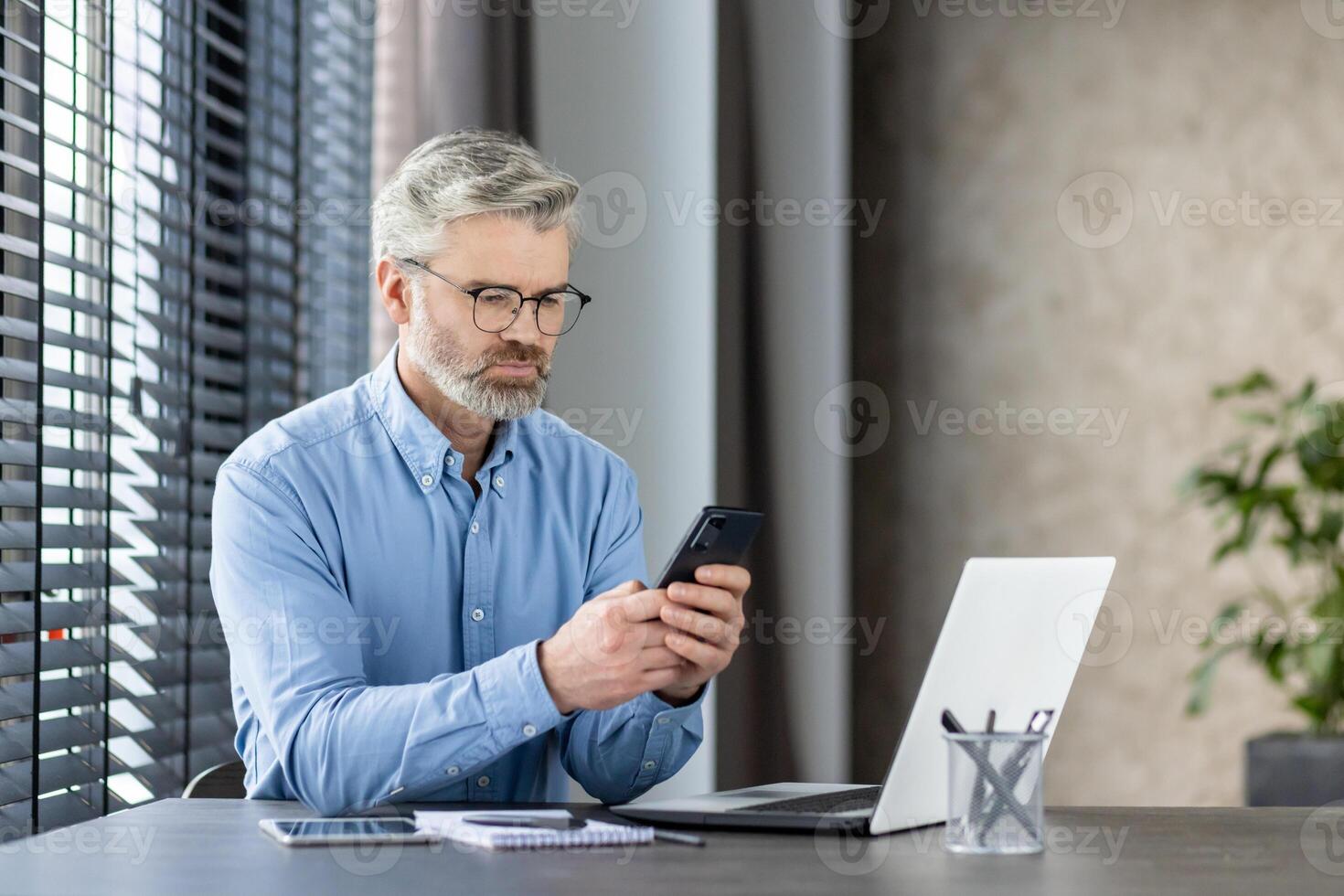 Mature businessman sitting at a desk, attentively using a mobile phone and working on a laptop in a modern office environment. photo