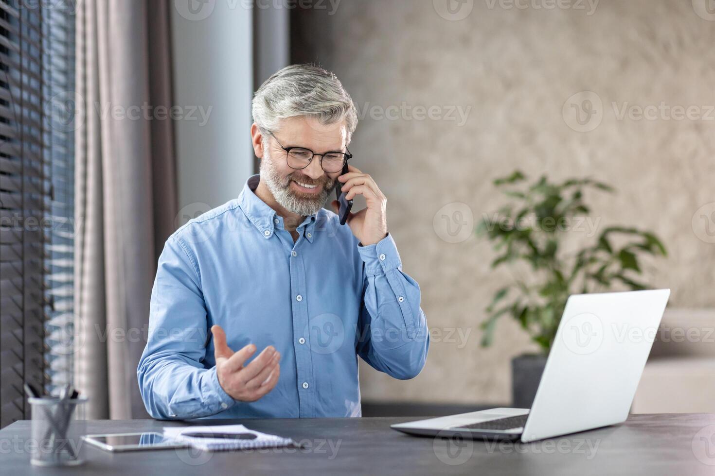 Smiling mature businessman communicating on phone while working on laptop at desk in a modern office with natural light and greenery in the background. photo