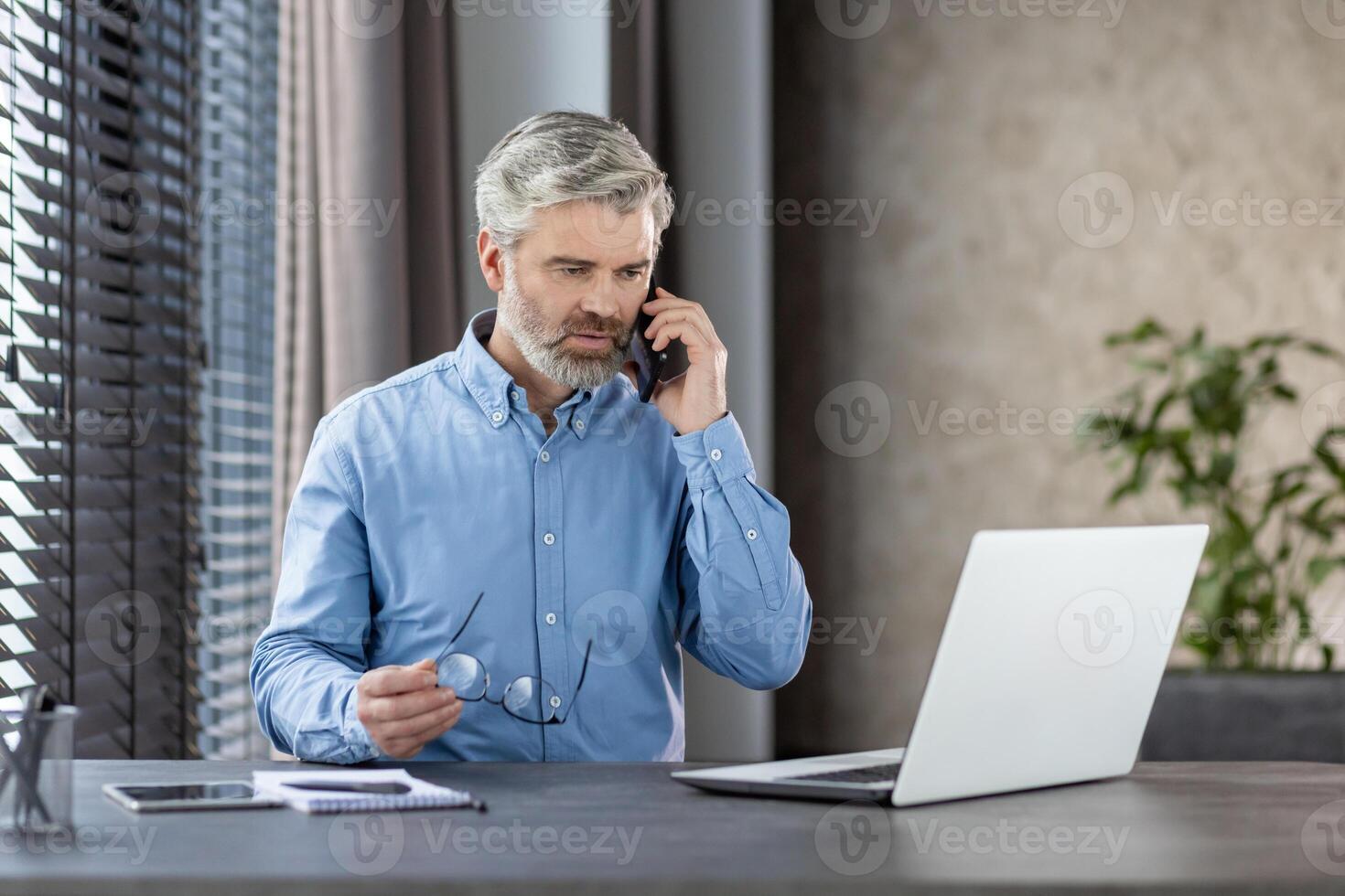 Mature businessman in blue shirt talking on phone while working on laptop at office desk. Professional working environment with natural light. photo