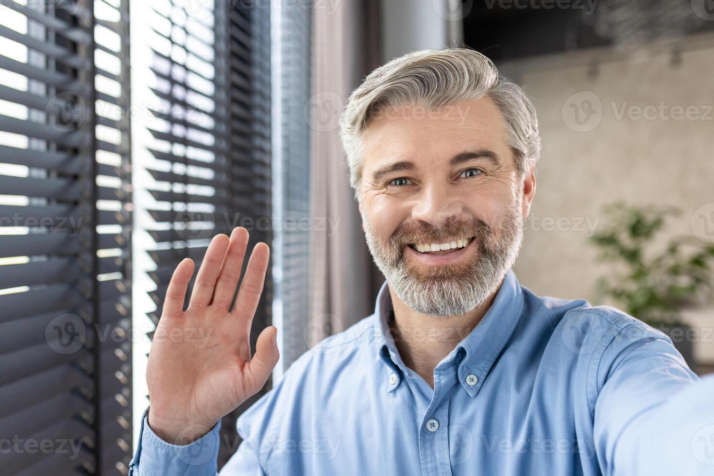 Happy man with gray hair and beard waving at the camera while having a call in a well-lit modern office. He is wearing a blue shirt and smiling warmly. photo