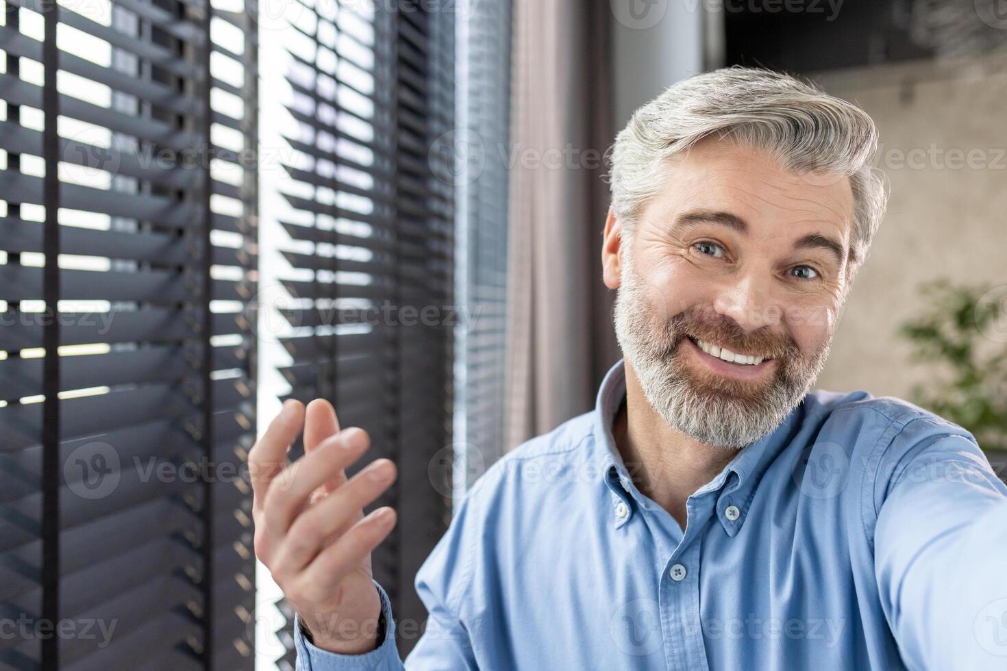 Smiling man with grey hair and beard having a call in bright office with blinds in the background. photo