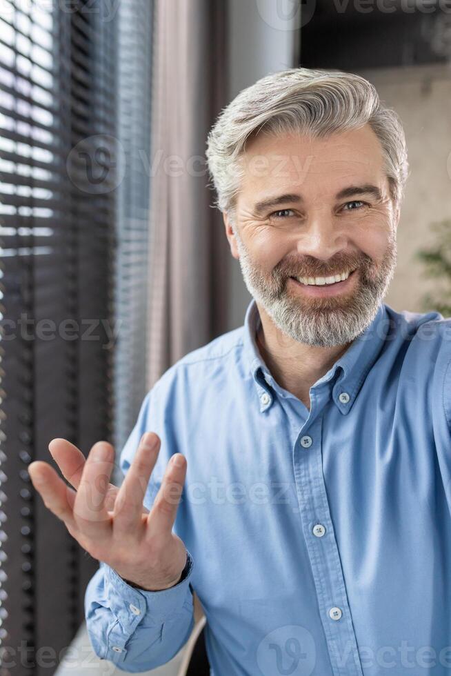 Smiling mature man in blue shirt having call using phone from office or home, engaging in conversation, looking at camera, and showing positive emotions photo