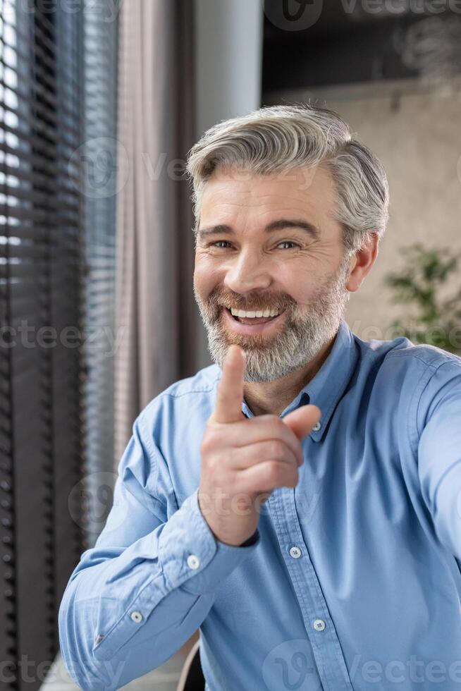 Middle-aged man in blue shirt smiling and pointing while calling from home office, expressing happiness and engagement. photo