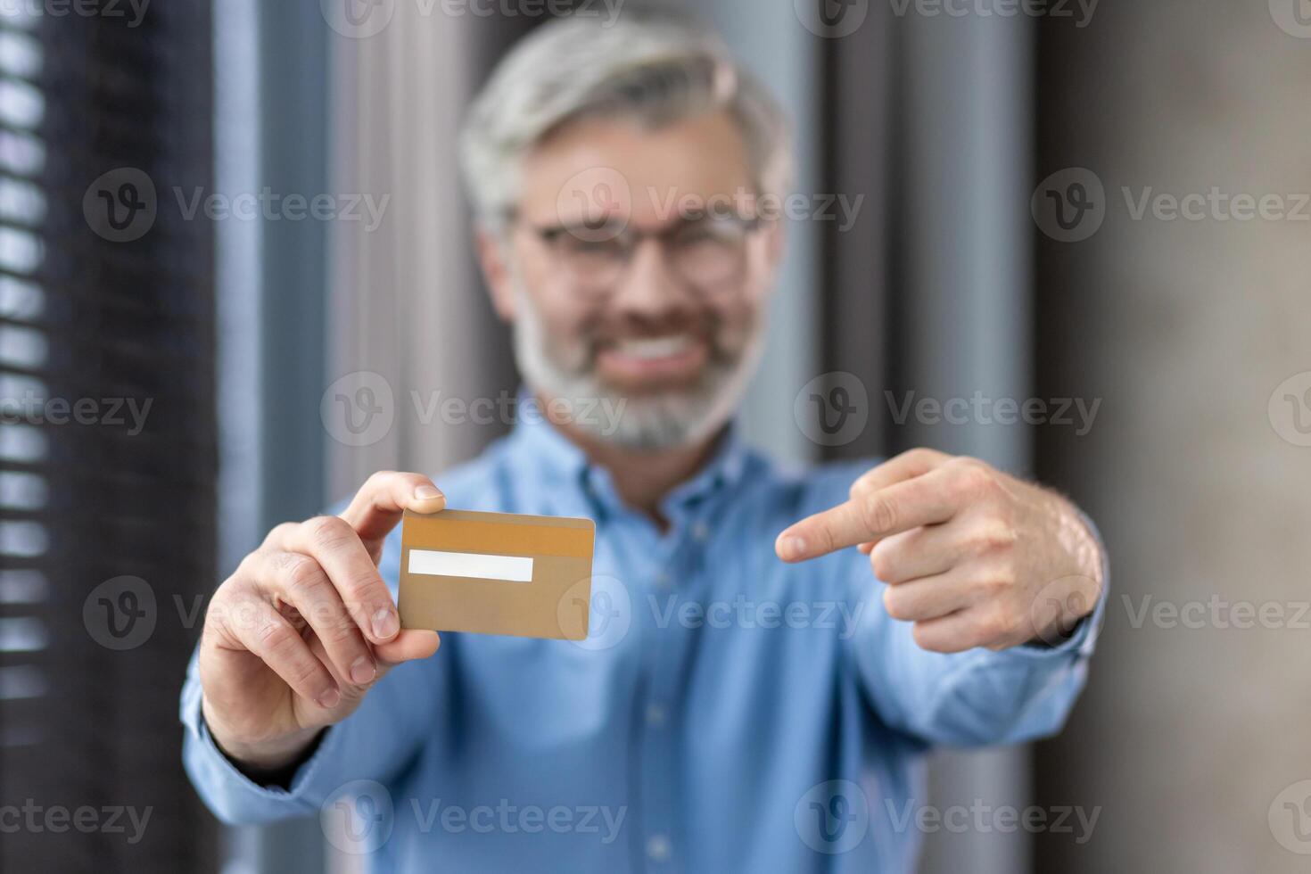 Middle-aged man with beard and glasses smiling, holding and pointing at a credit card, indoors photo