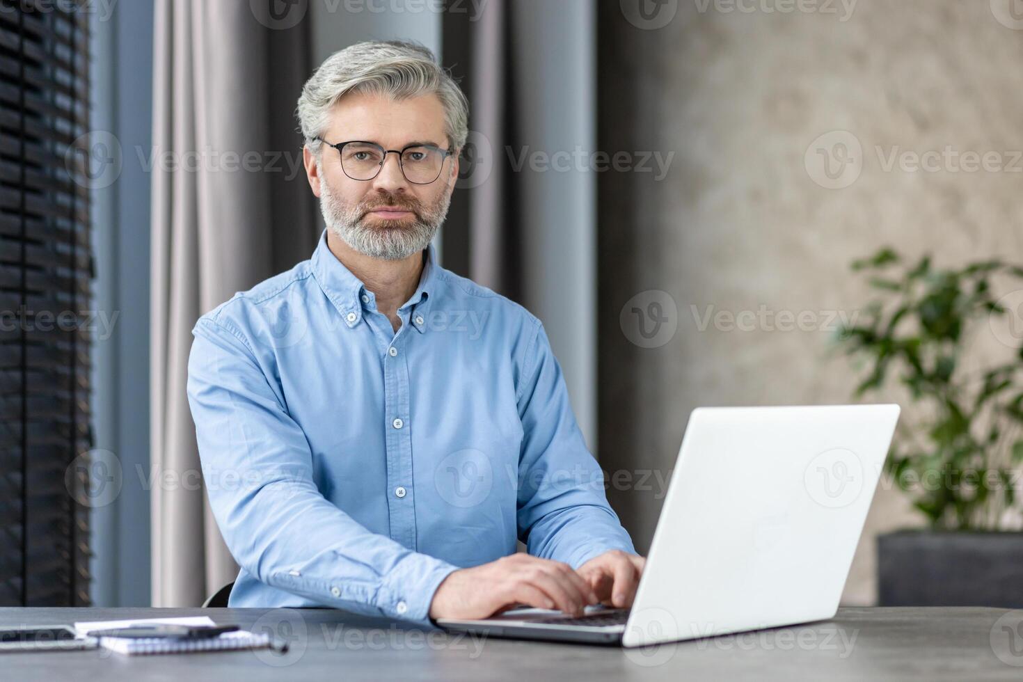 Confident mature businessman with glasses typing on a laptop in a modern office setting. The image portrays professionalism, focus, and a contemporary work environment. photo