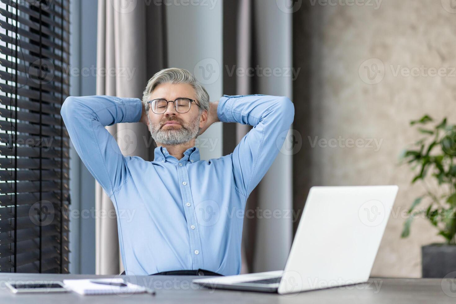 Mature businessman with glasses and beard taking a moment to relax at office desk. Hands behind head, laptop in front, serene expression. Concept of rest, leisure, workplace, and stress relief. photo