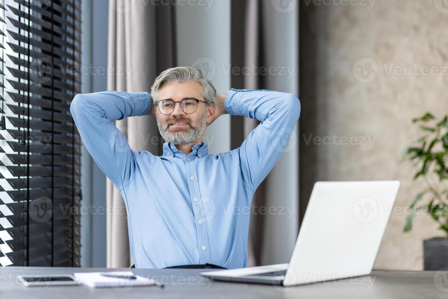 Mature male office worker taking a break with hands behind head, sitting at desk with laptop in a modern office. The man is wearing glasses and a blue shirt, looking content and relaxed. photo