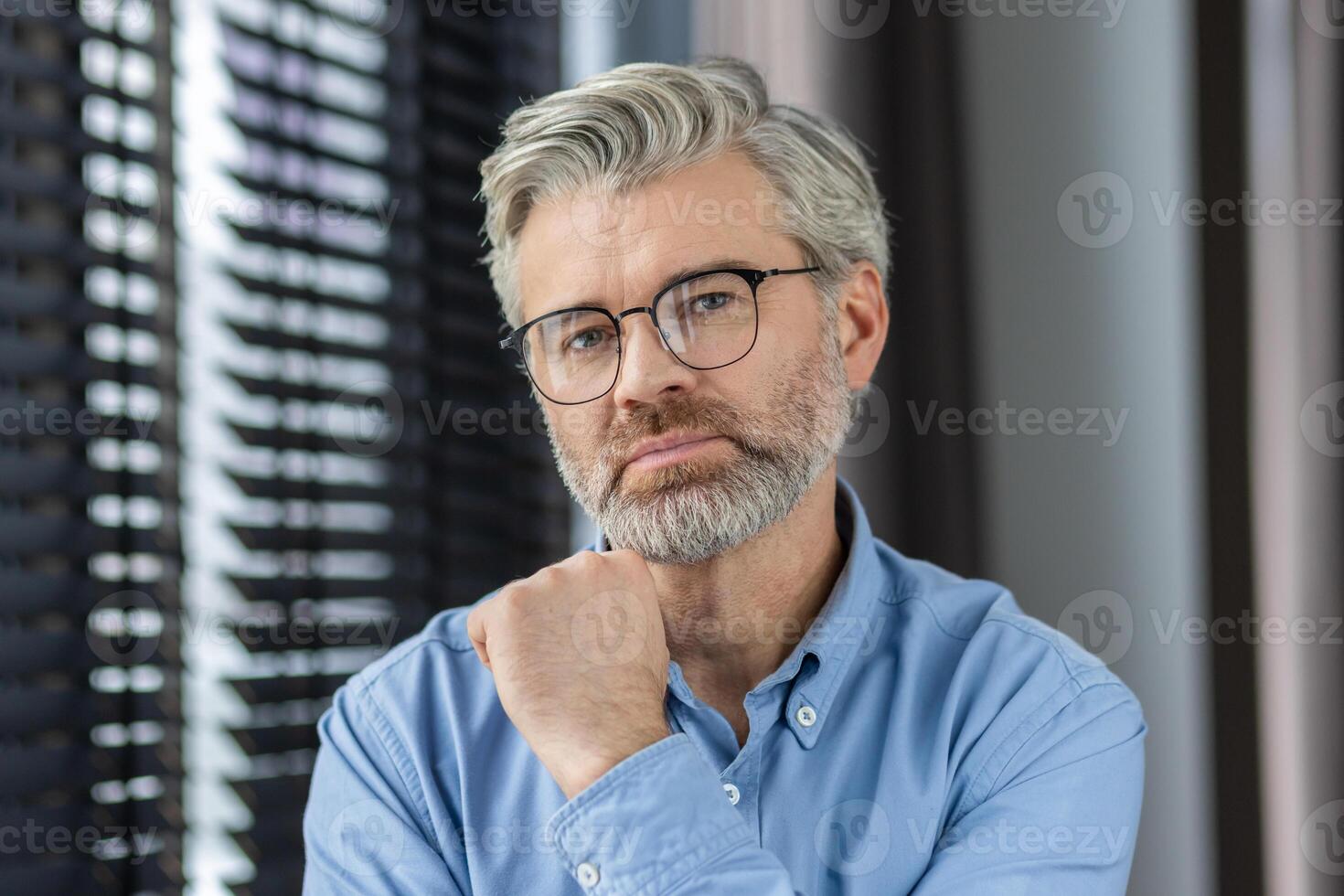 Confident mature man with glasses in a blue shirt posing indoors by window blinds, showcasing a thoughtful and reflective expression. photo