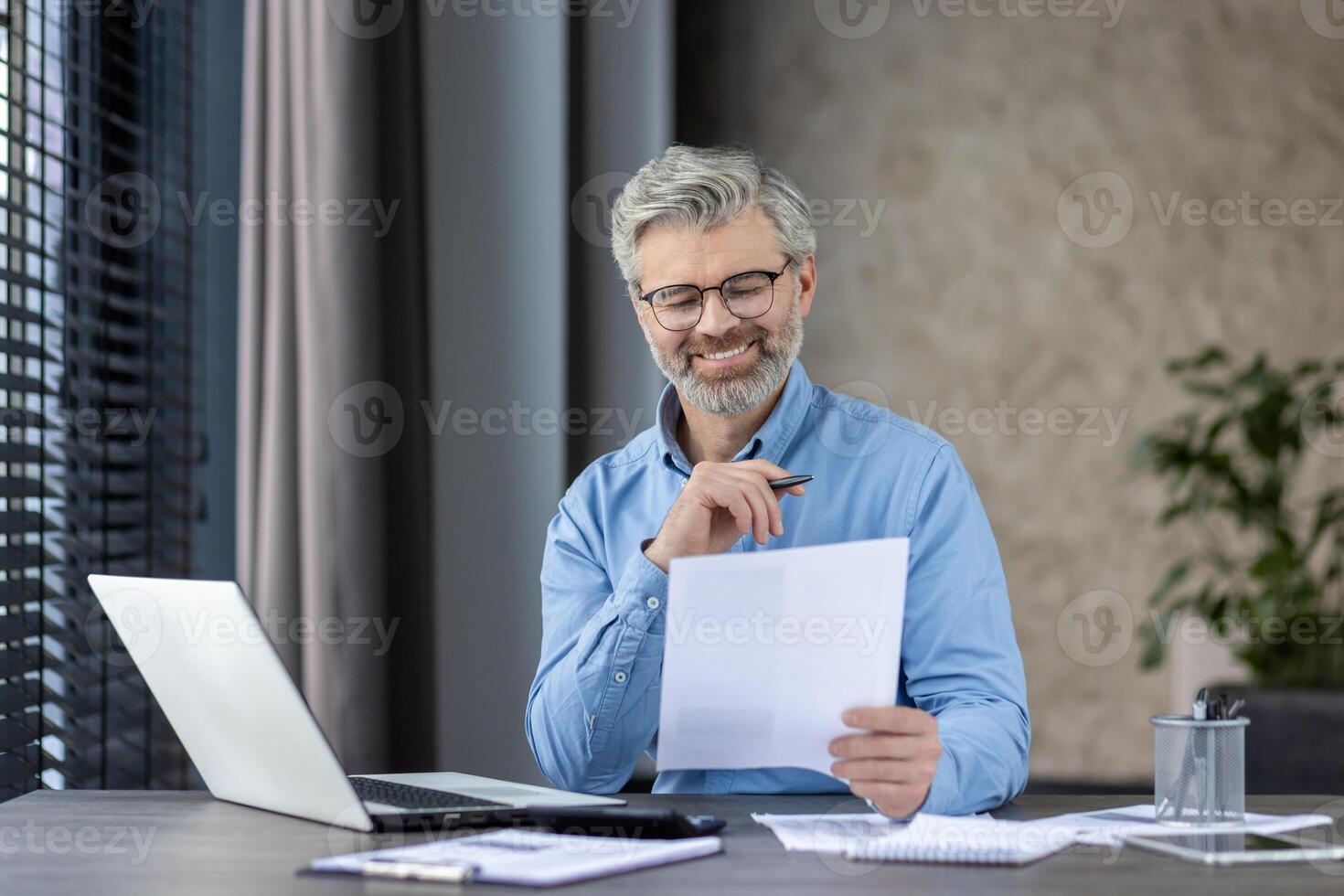 A smiling middle-aged businessman with glasses and beard reviews documents at his office desk. He is working on a laptop and appears content in a modern workspace. photo