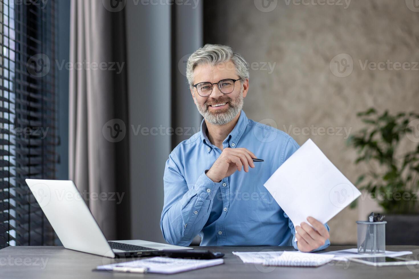 Mature businessman working at desk with laptop and documents in modern office, smiling confidently. Professional workspace with natural light. Successful and productive work environment photo