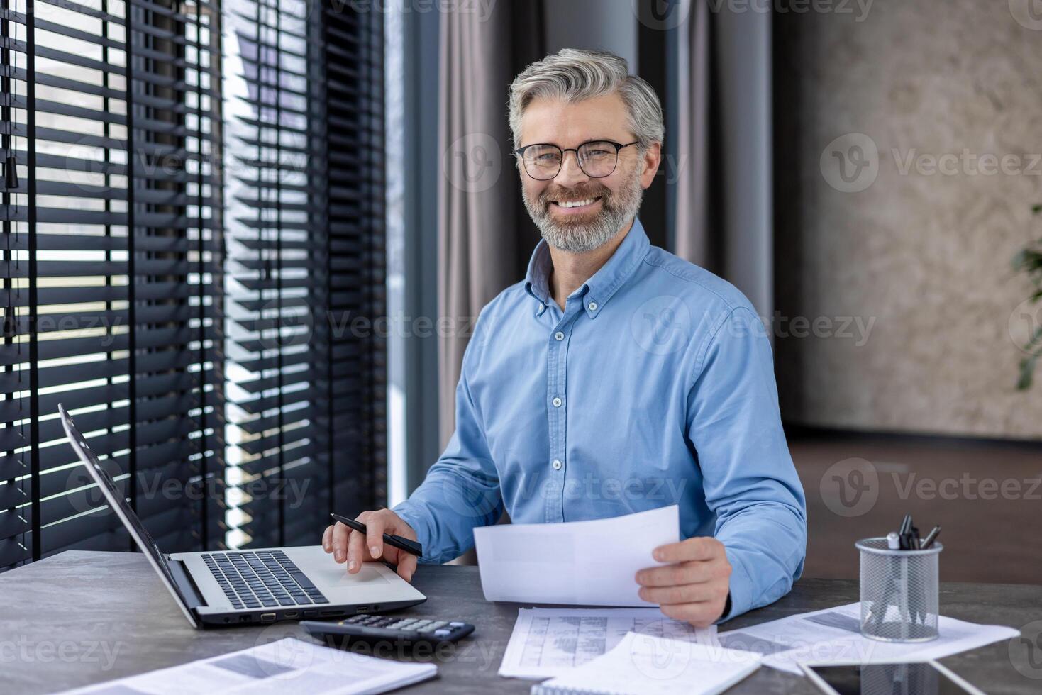 Smiling mature businessman working on a laptop while sitting at his desk in a modern office environment. Holding papers and surrounded by documents. photo